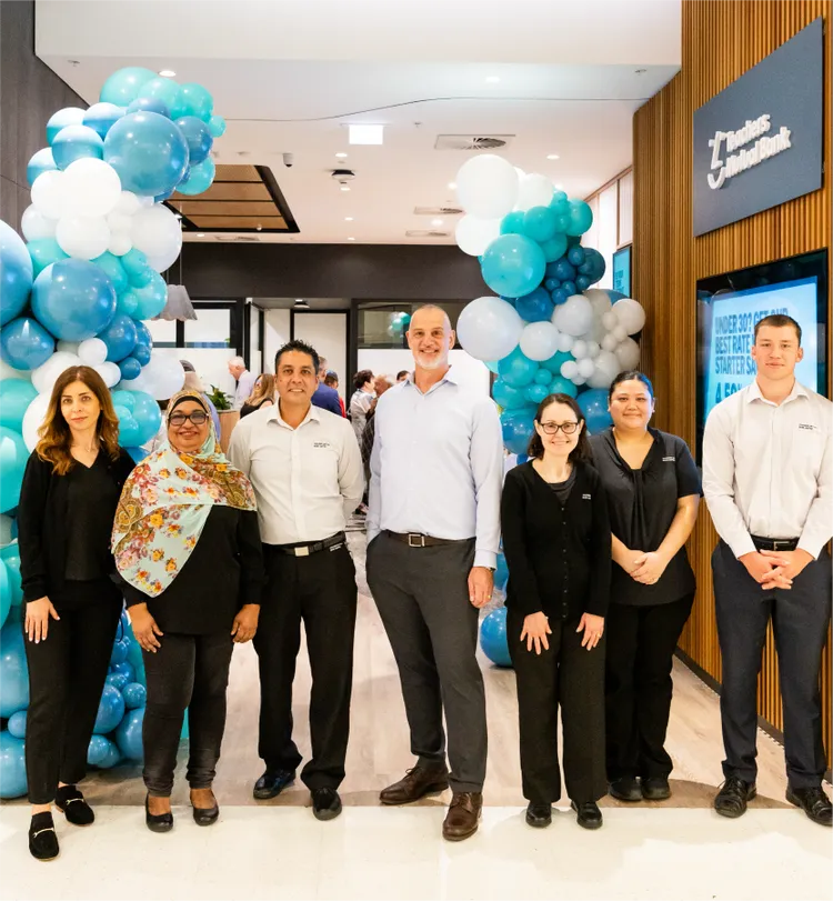 A group of adults stands in front of a business entrance decorated with blue and white balloon arches.