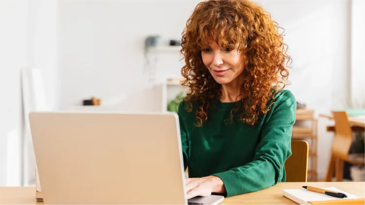 A woman with curly red hair sits at a wooden desk, working on a laptop.