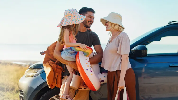 A family of three stands next to a blue car, preparing for a day at the beach.